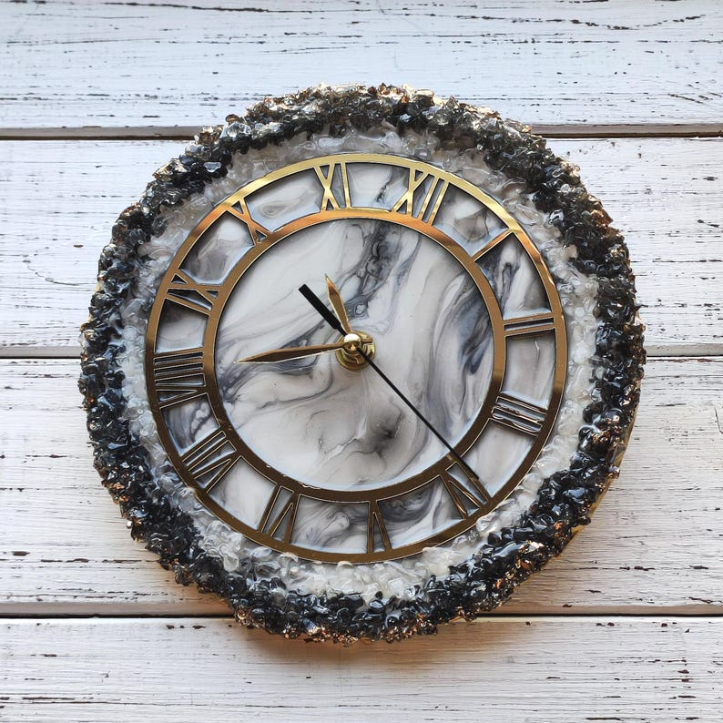 May include: Circular wall clock featuring a gold-coloured Roman numeral face and hands. The clock face has a white and grey marble design. The clock is framed by a textured, dark grey and black border. The clock is on a white wooden surface.