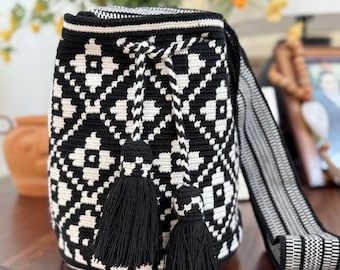 May include: Black and white crocheted shoulder bag with a geometric pattern. The bag features two black tassels and a matching strap. The bag is sitting on a wooden surface.