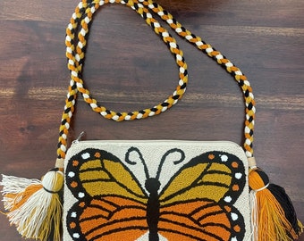 May include: A white beaded crossbody bag with a butterfly design. The butterfly is orange, yellow, and black. The bag has a braided strap with black, white, and orange accents and tassels.