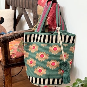 May include: A green and cream-coloured tote bag with a floral pattern and black and cream stripes. The bag has green straps with pink stripes and a green tassel. The bag is sitting on a wooden chair.