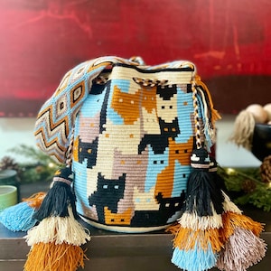 May include: A handmade bucket bag featuring a repeating cat pattern in black, tan, light blue, and cream. The bag has a patterned shoulder strap and decorative tassels in coordinating colors. The bag is made of woven fabric.
