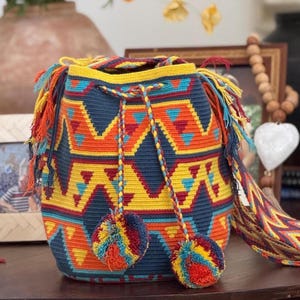 May include: A hand-woven bag with a geometric pattern in navy blue, yellow, orange, and turquoise. The bag has a drawstring closure, colourful tassels, and two pom-poms. A unique accessory.