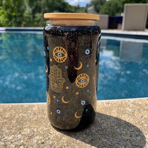 May include: A black glass tumbler with a bamboo lid, decorated with gold and blue celestial designs, including eyes, moons, stars, and a Hamsa hand. The tumbler is cylindrical and appears to be filled with a dark liquid.
