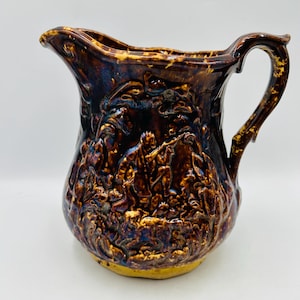 May include: A brown ceramic pitcher with a detailed relief design. The pitcher features a handle and a spout. The relief depicts figures in a landscape. The pitcher has a glossy finish with hints of blue and gold.