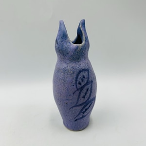 May include: A handcrafted ceramic vase in a soft lavender hue, featuring a unique, elongated shape with two upward-curving spouts. The vase is adorned with a dark blue leaf-like design. The speckled texture adds to its rustic charm.