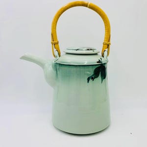 May include: A light green ceramic teapot with a bamboo handle. The teapot has a spout and a lid. The handle is a light brown color. The teapot has a painted leaf design on the side.