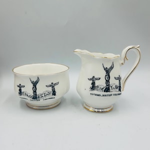 May include: A white ceramic creamer and sugar bowl set. Each piece features a black and white illustration of totem poles with the text "TOTEMS, BRITISH COLUMBIA" in black. The rims and bases are trimmed with gold.
