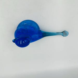 Puede incluir: Una figura de caracol de cristal azul vibrante. El caracol tiene un cuerpo redondeado, una concha en espiral y una antena larga y delgada. El cristal es de un azul profundo y transparente, y la figura est&aacute; sobre un fondo blanco.