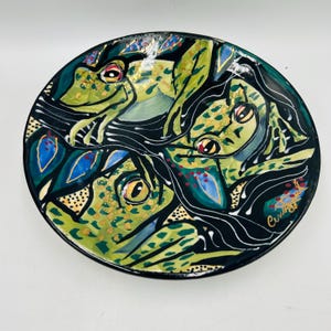 May include: A decorative ceramic plate featuring a hand-painted design of three green frogs with yellow eyes and red accents. The frogs are set against a black background with stylized leaves in blue and gold. The plate has a glossy finish.