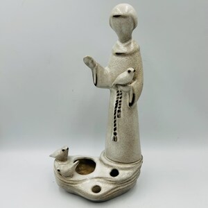 May include: A white ceramic figurine of Saint Francis of Assisi holding a bird. The figurine is standing on a base with three candle holders and two small birds.