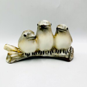May include: A ceramic figurine featuring three small birds perched side-by-side on a textured branch. The birds have cream-colored bodies with grey-brown shading on their heads and wings, and dark brown painted details for their eyes and feet.