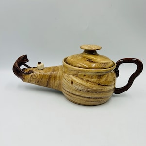 May include: A ceramic teapot with a brown handle and lid, featuring a brown cat figurine on the spout. The teapot has a beige and brown marbled design, with a small white object near the cat.