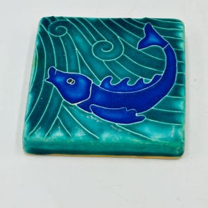 May include: Square ceramic tile with a blue fish design. The fish is a deep blue color and is set against a teal background with wavy lines. The tile has a glossy finish and a yellow edge.