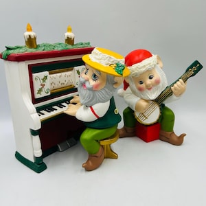 May include: A whimsical Christmas figurine set featuring two gnomes. One gnome plays a white, red, and green piano, while the other strums a banjo. The piano has two candle lights on top. The gnomes wear festive hats and green outfits.