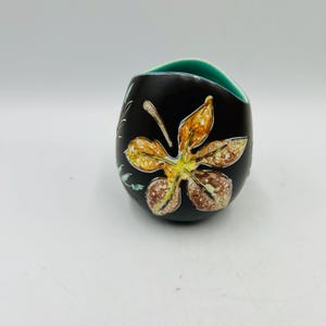 Puede incluir: Un pequeño cuenco de cerámica negro con un borde turquesa y un diseño pintado a mano de una hoja amarilla y marrón. El cuenco es ligeramente ovalado y tiene un diseño único y asimétrico.