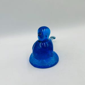 Puede incluir: Una figura de vidrio azul cobalto de una figura sentada con un cuerpo redondeado y un peque&ntilde;o ap&eacute;ndice curvado. La figura se asienta sobre una base circular texturizada. El vidrio tiene un acabado brillante.