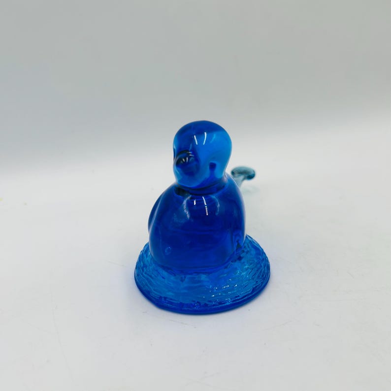 Puede incluir: Una figura de vidrio azul cobalto de una figura sentada con un cuerpo redondeado y un peque&ntilde;o ap&eacute;ndice curvado. La figura se asienta sobre una base circular texturizada. El vidrio tiene un acabado brillante.