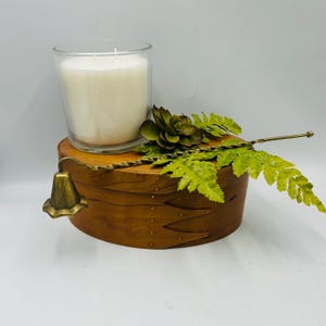 May include: A candle in a clear glass jar sits atop a wooden box with a decorative brass bell and faux greenery. The candle is white, and the box is a warm brown color. The greenery includes a succulent and fern-like leaves.