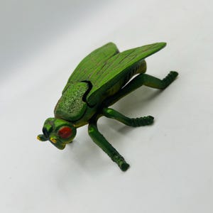 May include: A green plastic insect toy with red eyes and detailed wings. The toy insect has six legs and is positioned on a white surface. The insect's body is a darker green than its wings.