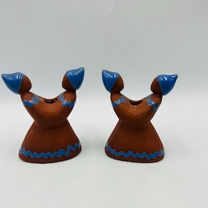 May include: Two ceramic candle holders in a Native American design. The candle holders are brown with blue accents, including a wavy line around the base and blue headpieces. The candle holders are on a white background.