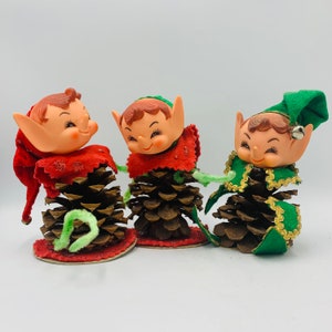 May include: Three Christmas elf ornaments made of pine cones and plastic. Each elf has a different colored hat, red, green, and green with gold trim. The elves have smiling faces and are wearing a red, green, and green with gold trim fabric.