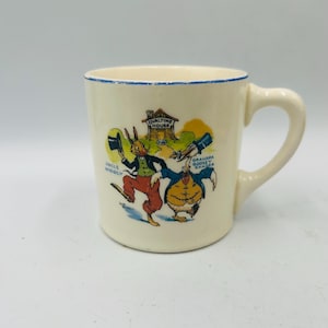 May include: Cream-colored ceramic mug with a blue rim and a curved handle. The mug features a colorful illustration of Uncle Wiggily and Grandpa Goosey Gander. The text "Ovaltine House" is also visible.