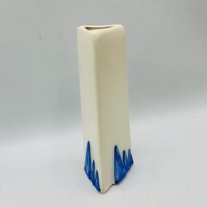 May include: A tall, rectangular vase with a cream-colored body and a triangular opening. The base features a series of blue, stylized mountain-like shapes. The vase is a decorative item, suitable for displaying flowers or as a standalone art piece.