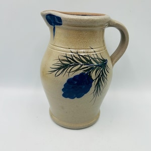 May include: Tan stoneware pitcher with a handle and spout. The pitcher is decorated with a blue pine cone and green pine needles. The rim of the pitcher is a darker brown color.