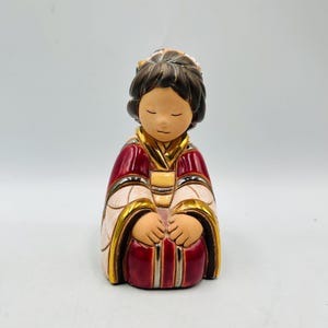 May include: A ceramic figurine of a seated person with closed eyes. The figure wears a red and gold robe with white accents. The hair is dark brown, and the skin tone is light. The figurine has a serene expression and is likely a decorative item.