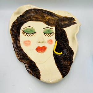 May include: A ceramic wall plate with a stylized image of a woman's face. The woman has closed eyes, red lips, and a yellow earring. The plate is white with brown and green accents.