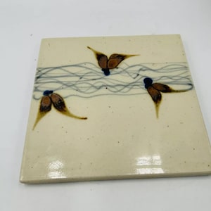May include: A square ceramic tile with a white background and a blue and brown abstract design. The design features a series of thin, wavy lines in blue and three brown butterfly-like shapes.