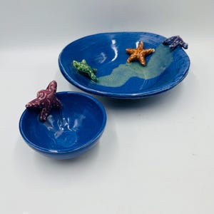 May include: Two blue ceramic bowls with starfish accents. The larger bowl has a wave design with a green, orange, and purple starfish. The smaller bowl has a purple starfish. The bowls are on a white surface.