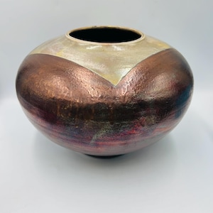 May include: A round ceramic vase with a wide opening. The top section is a light beige color, with a copper-colored band below. The bottom of the vase has a reddish-brown hue.