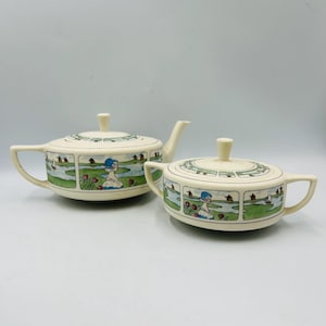 May include: Cream-colored ceramic teapot and sugar bowl set. Each piece features a hand-painted landscape scene with a woman in a blue hat, windmills, and a river. The teapot has a curved handle and spout, while the sugar bowl has two handles and a lid.
