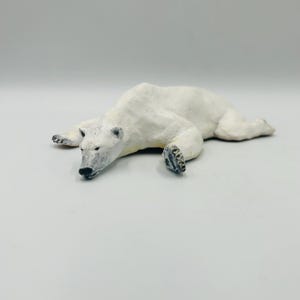 May include: A white polar bear figurine lying on its belly with its head turned to the side. The bear has black eyes and a black nose.