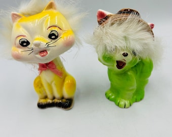May include: Two vintage ceramic cat figurines. One is yellow with a pink bow and the other is green. Both have white fur around their faces. The cats have black eyes and are smiling.
