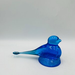 Puede incluir: Una figura de p&aacute;jaro de cristal azul vibrante. El p&aacute;jaro tiene un cuerpo redondeado, una cabeza peque&ntilde;a y una cola larga y delgada. La base del p&aacute;jaro es texturizada y el cristal tiene un acabado brillante. Es un art&iacute;culo decorativo.