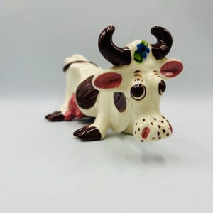 May include: A ceramic cow figurine with brown and white spots. The cow has pink ears, brown horns, and a flower on its head. The cow is lying down with its head tilted to the side.