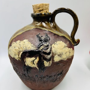 May include: A brown ceramic jug with a cork stopper and a handle. The jug has a painted design of a black and white animal with white clouds in the background.