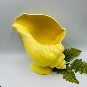 May include: A bright yellow ceramic seashell-shaped vase. The vase has a glossy finish and sits on a small, round base. The opening of the vase is wide and curved. A green fern branch is next to the vase.