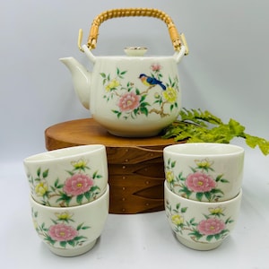 May include: A cream-colored ceramic tea set featuring a teapot and four teacups. The teapot has a woven handle and is decorated with a bird and floral design in pink, yellow, and blue. The teacups have a matching floral pattern.