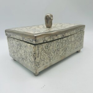 May include: A rectangular decorative box with a lid. The box has a textured, off-white surface with dark gray veining. The edges and feet are silver-colored. A small, silver-colored handle is on top of the lid.