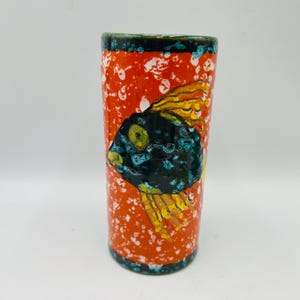 Puede incluir: Un jarrón de cerámica cilíndrico con fondo naranja brillante y un patrón de salpicaduras blancas. Un pez negro con aletas amarillas y ojos verdes está pintado en el jarrón.