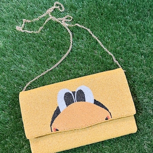 May include: Yellow beaded clutch with a cartoon duck design. The clutch features a gold chain strap. The duck has black eyes, a light blue head, and an orange beak. The clutch is rectangular and has a flap closure.