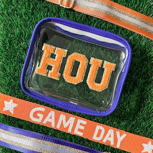 May include: Clear stadium bag with orange "HOU" letters and a purple trim. Includes orange and purple straps with silver accents and the text "GAME DAY". The bag and straps have silver metal hardware.