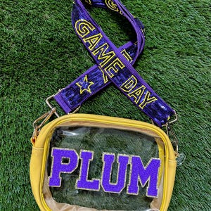 May include: A yellow crossbody bag with a clear front panel and the word "PLUM" in purple block letters. The bag has a purple and gold "GAME DAY" strap.