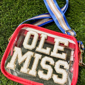 May include: Clear stadium-approved crossbody bag with the text "OLE MISS" in white and gold lettering. The bag has a red trim and a blue, white, and gold striped strap. The bag is on a green surface.