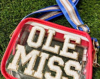 May include: Clear stadium-approved crossbody bag with the text "OLE MISS" in white and gold lettering. The bag has a red trim and a blue, white, and gold striped strap. The bag is on a green surface.