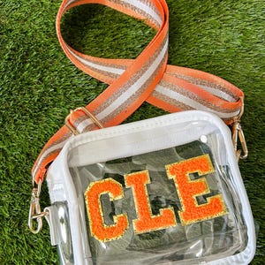May include: A clear crossbody bag with a white trim and an orange and gold strap. The bag features the letters "CLE" in orange with a gold outline. The strap is orange with white and gold stripes and gold hardware.