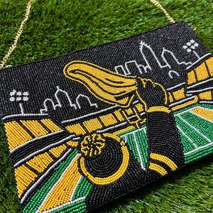 May include: A rectangular beaded clutch bag featuring a black, yellow, and green design of a football stadium scene with a city skyline in the background. The bag includes a gold-tone metal chain strap and depicts a figure waving a yellow towel.